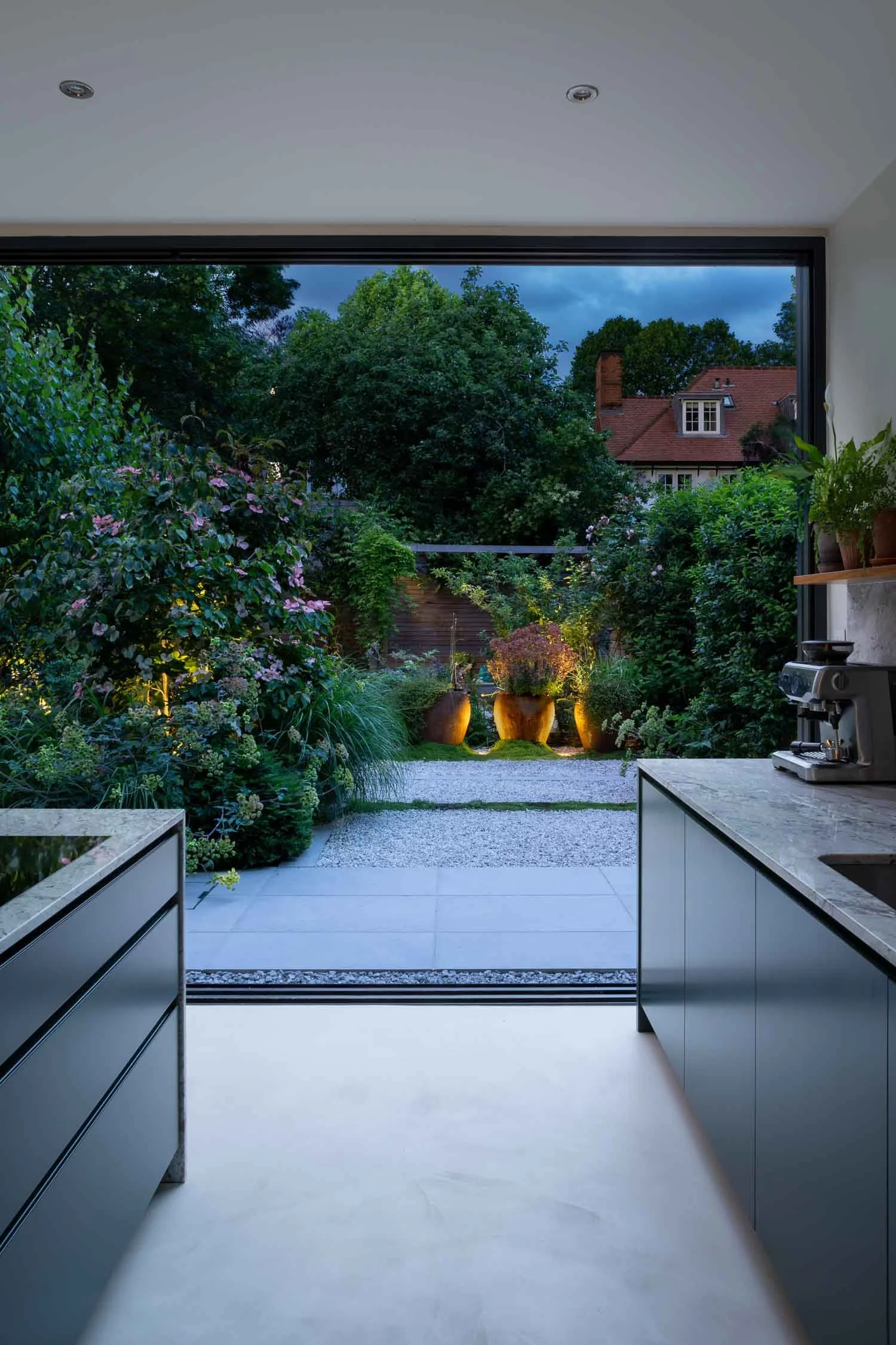 Open plan kitchen leading to densely planted town garden with low lighting at night