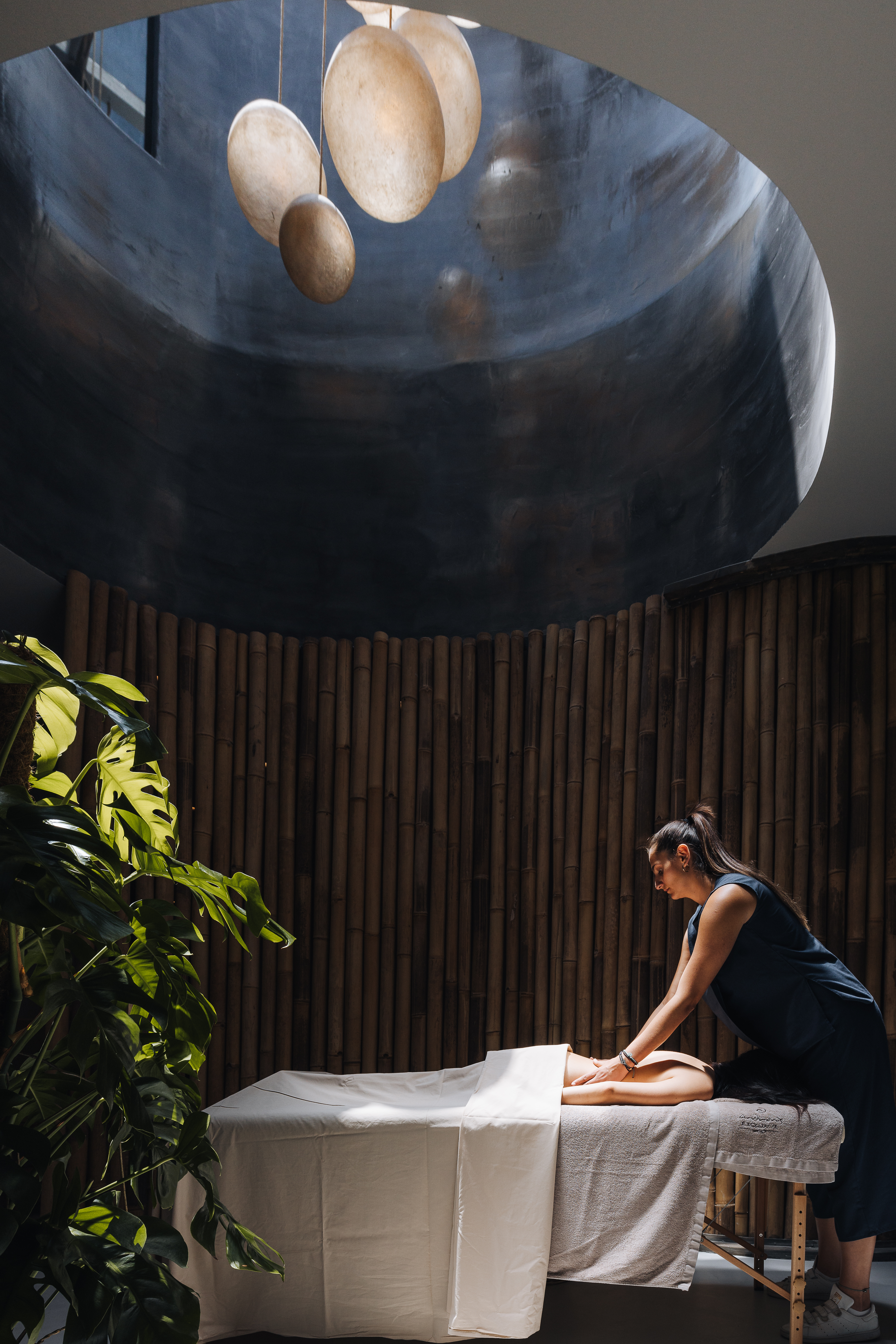 A female member of a spa administrating a treatment massage on a woman in a wood-clad room filled with natural light.