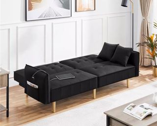 A black sofa bed with wooden legs opened up into a bed, in a living room with hardwood floors, a coffee table, a lamp, and art on the walls.