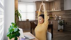 woman in a yellow top sitting in front of a laptop with arms overhead stretching. there's a brown kitchen behind her