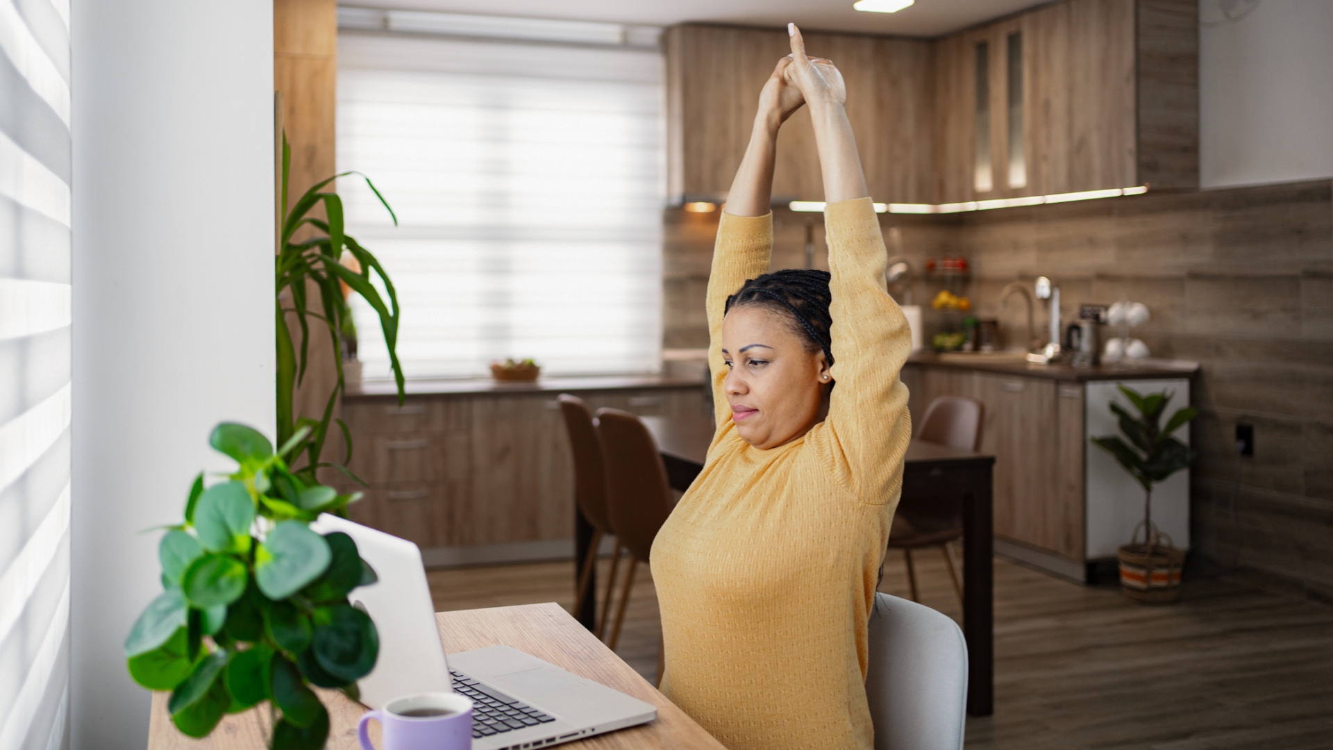 woman in a yellow top sitting in front of a laptop with arms overhead stretching. there&#039;s a brown kitchen behind her 