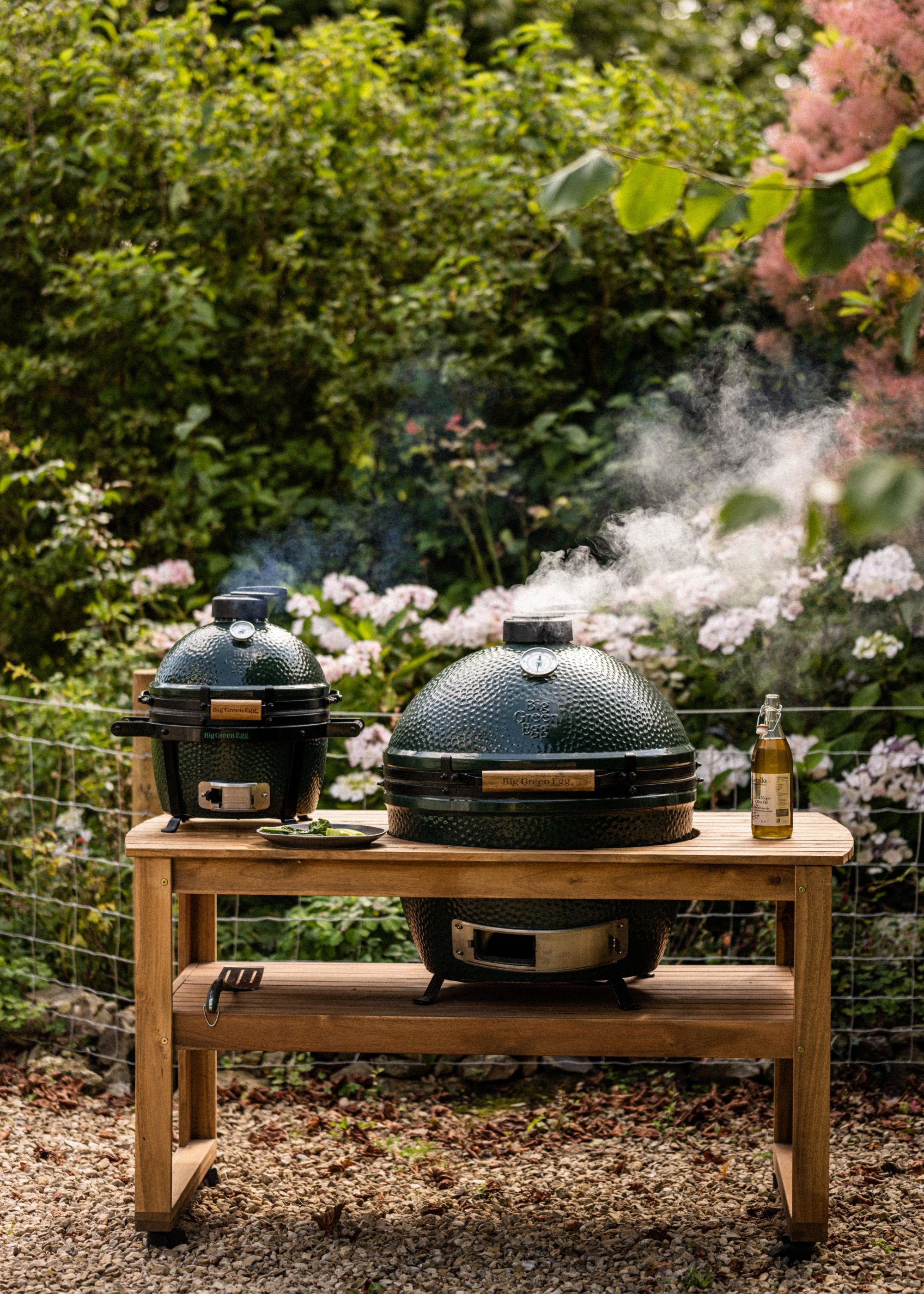 Two small and large Big Green Eggs in a garden with lots of plants and flowers around it