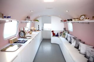 Interior of a bus with pastel decor, white sofa, canvas throw cushions and objects on white shelves against pink walls