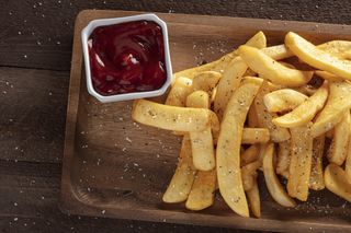 A portion of chips served on a wooden serving board with seasonings and a side portion of tomato ketchup.