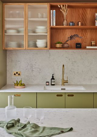 Fluted glass cabinet in a kitchen next to open shelf cabinets. At the bottom are sage green colored kitchen cabinets with a sink and gold tap hardware.