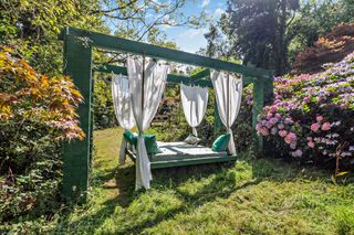 A swinging bed sits in the garden surrounded by a variety of plants such as big leaf hydrangeas and maple trees