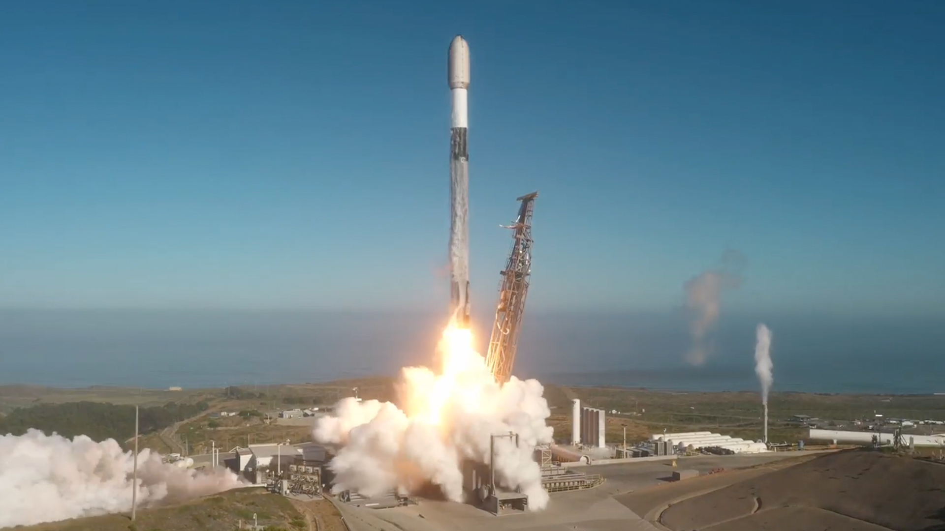 A white, black and gray rocket lifts off into a blue sky from an oceanfront launch pad.