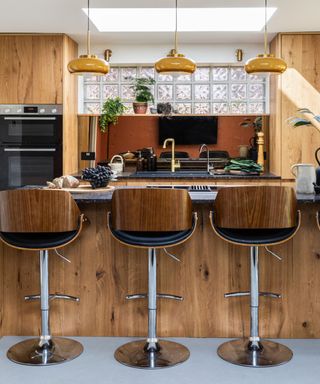 A kitchen island with a wooden base, black granite countertop with a wooden board on it, three curved wooden and metal bar stools, and three yellow glass pendant lights above it