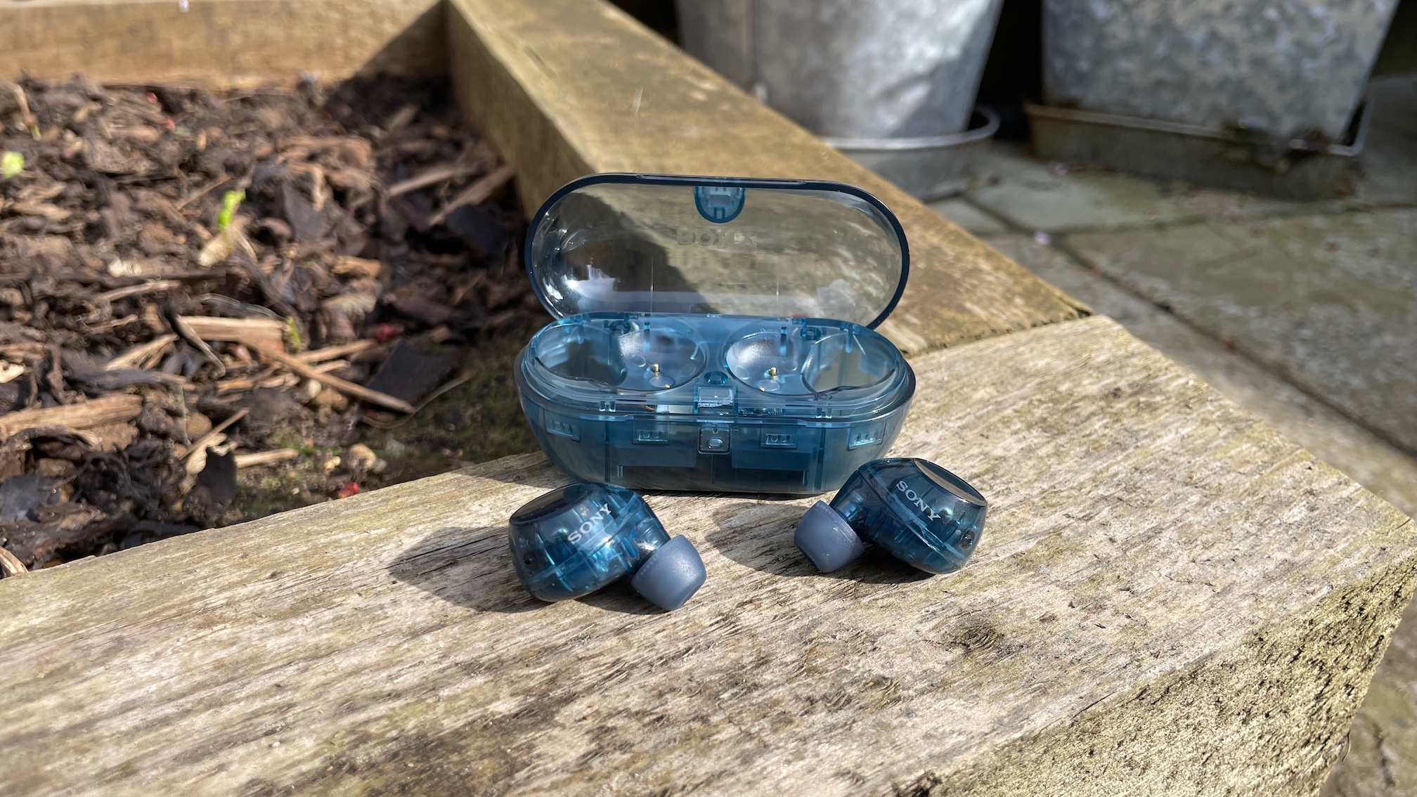 A blue pair of Sony WF-C710N wireless earbuds with their charging case on a wooden railway sleeper by a flower bed.