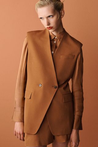 A female model wearing a brown jacket, a brown button up shirt and brown shorts.