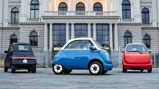 Three Microlino electric city cars on a plaza, in black, blue and red