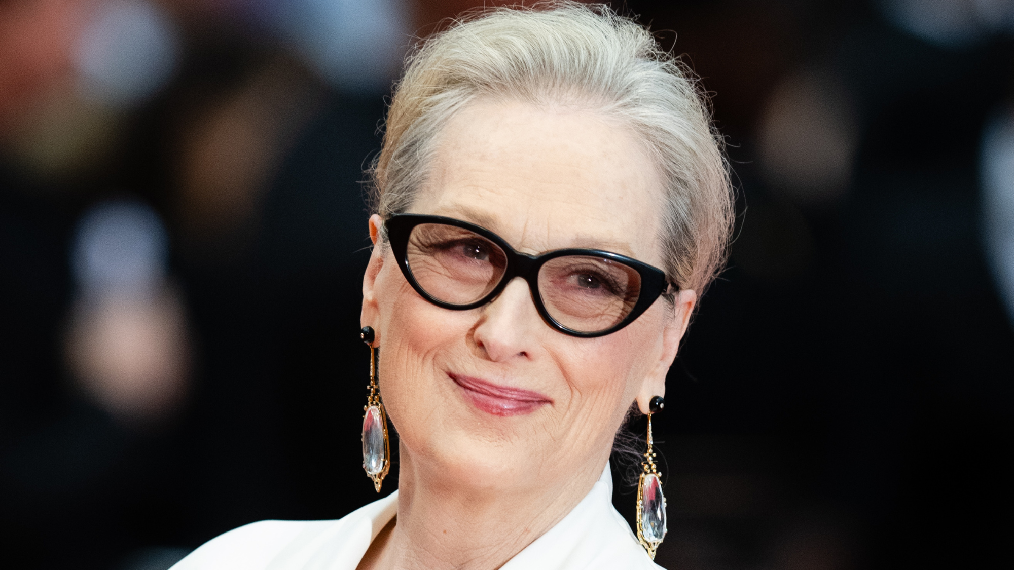 Meryl Streep wearing glasses and clear dangly earrings and smiling on a red carpet.