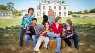 Sarah Beeny with her husband Gareth and their 4 sons sat in front of their white self build property and greenhouse in a large front garden