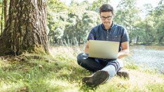 A man sitting beneath a tree by a river, using a laptop.