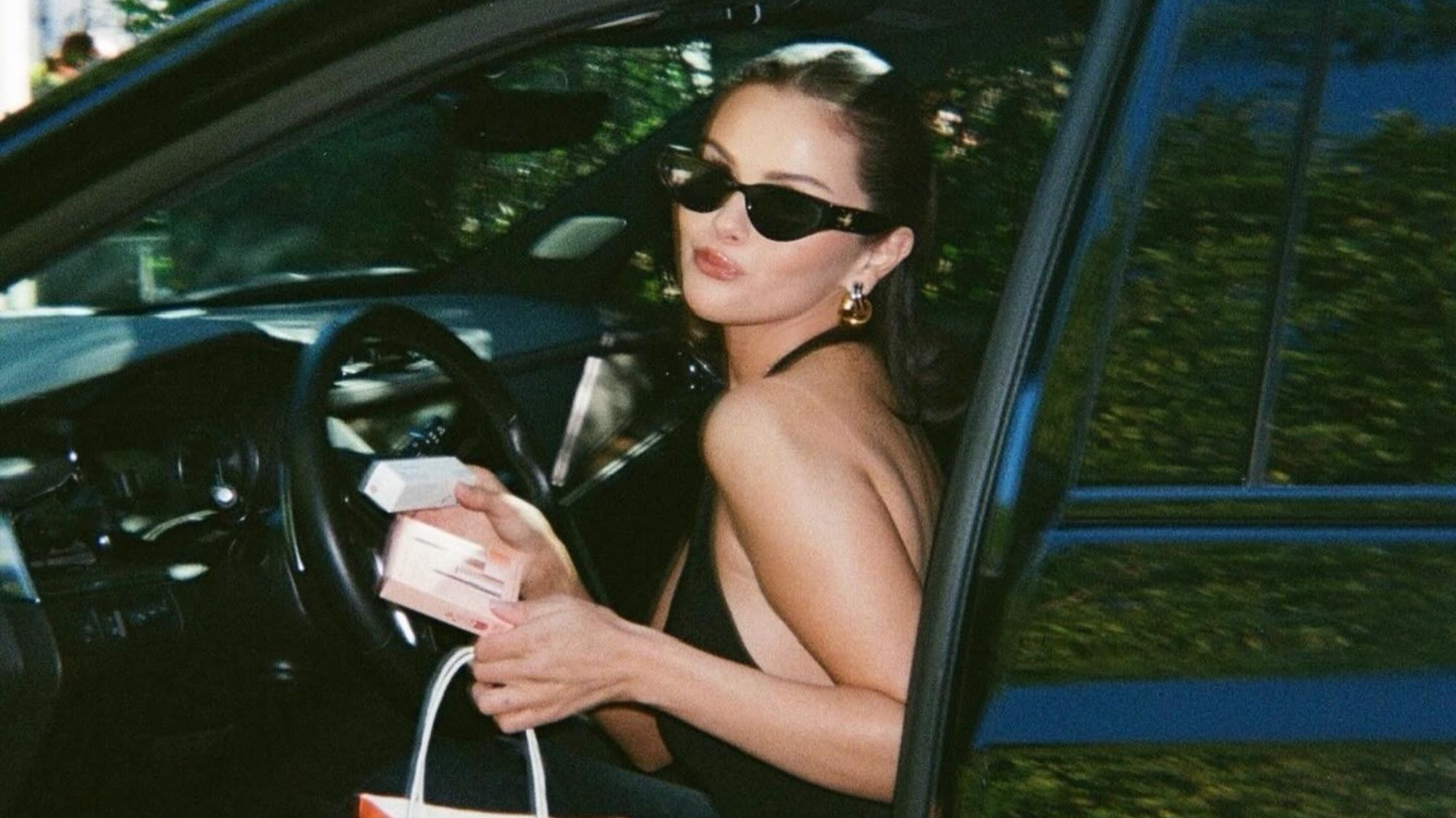 An image of Selena Gomez holding two Rare Beauty products and an Ulta shopping bag while posing in the open door of a car.
