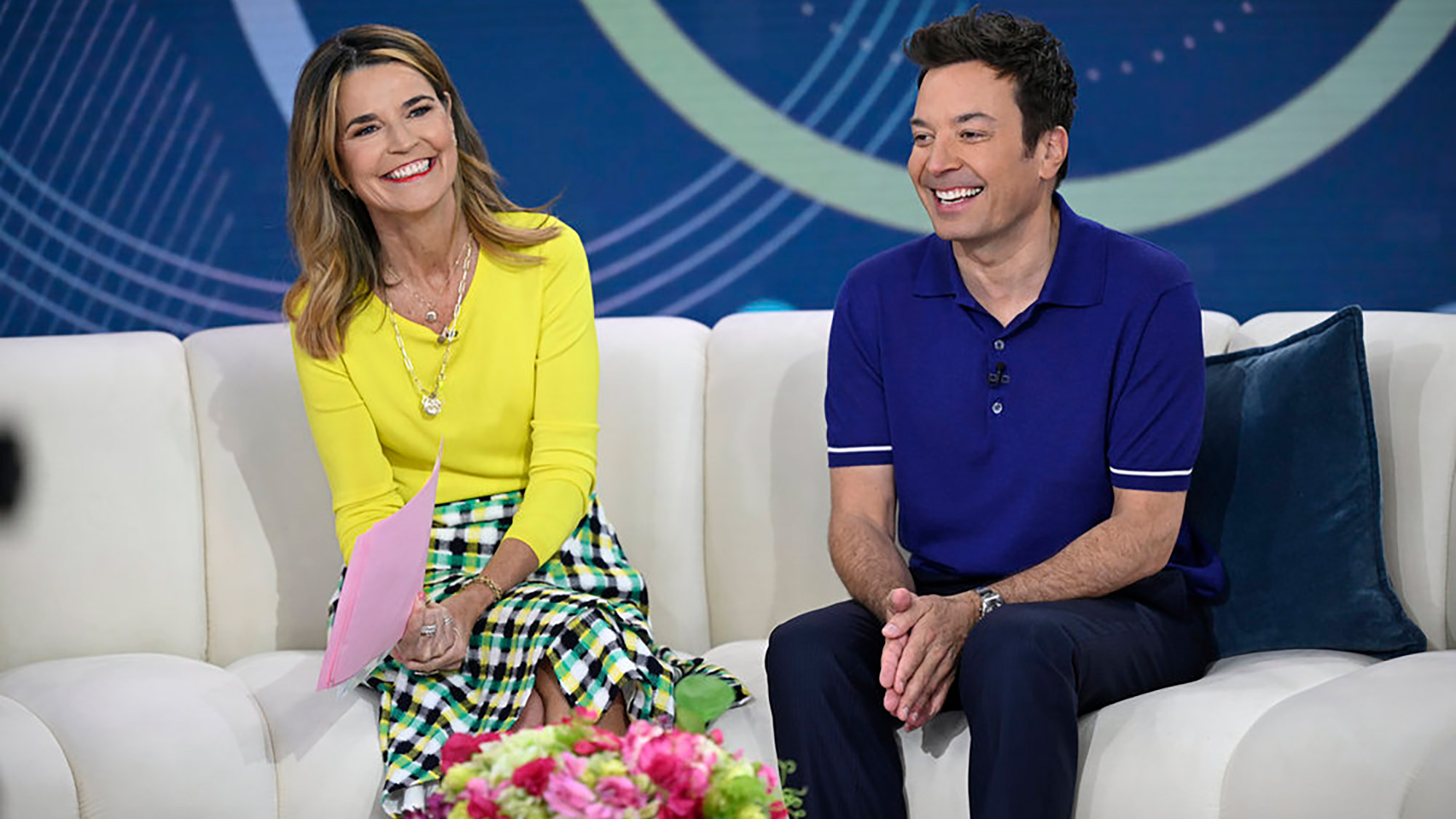 Jimmy Fallon promotes Wordle with Savannah Guthrie while sitting on the white couch on Today. She has a yellow top on and he is wearing a navy polo,