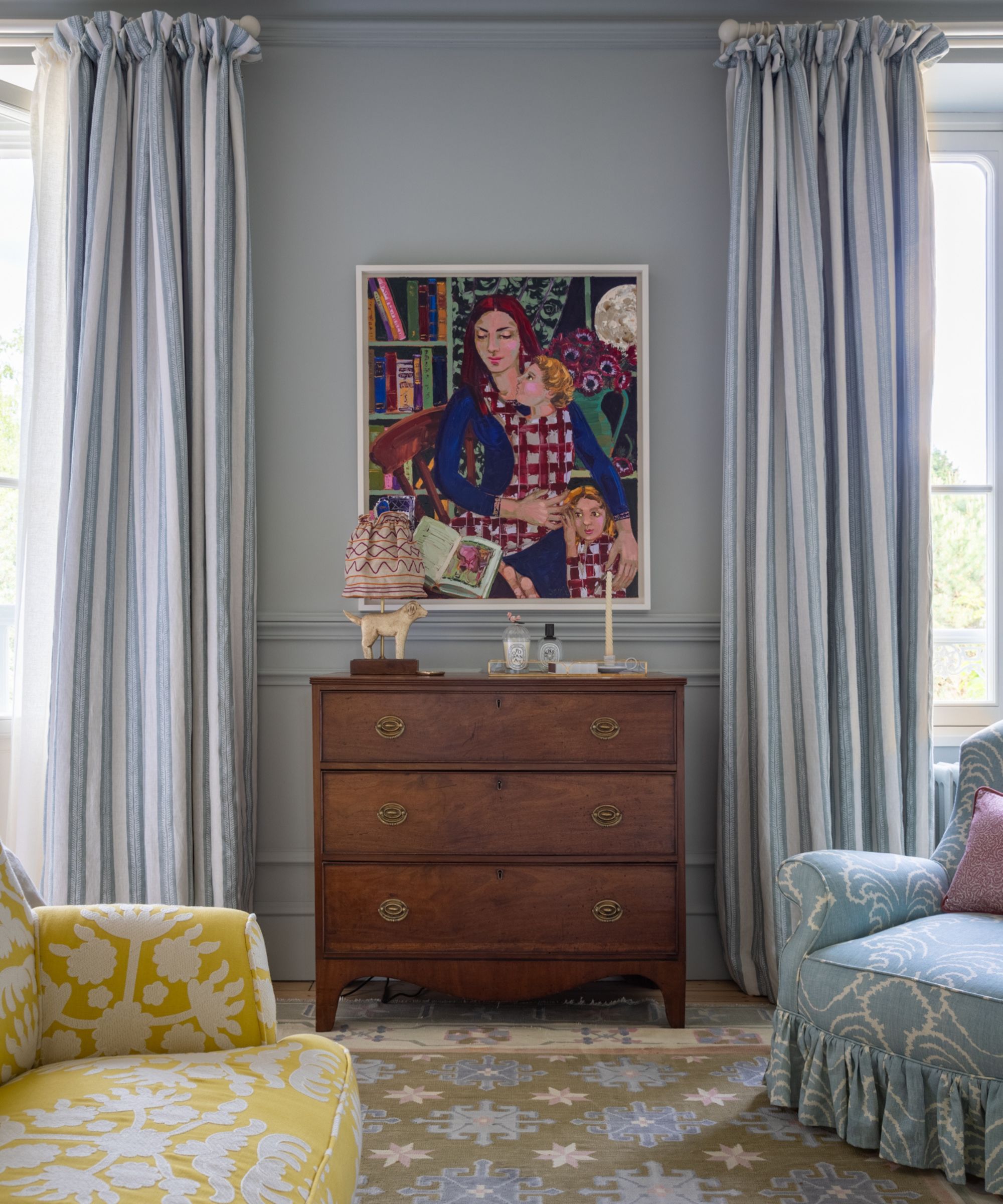 blue bedroom with blue and white striped drapes, a yellow foot of bed couch, a vintage chest of drawers and colorful painting