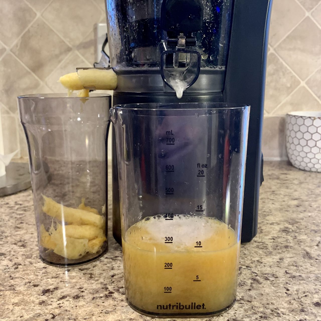 A close up of the base of a black plastic cold-press juicer, dispensing pale yellow fruit solids into a beaker on its left, and pale yellow juice into a clear beaker in front of it on a granite kitchen countertop.