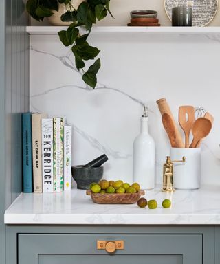 A kitchen corner with a marble panel, a white wall shelf with a trailing plant and plates, a white surfaces with cookbooks, a bowl of small green fruit and a container of wooden utensils, and a dark gray drawer