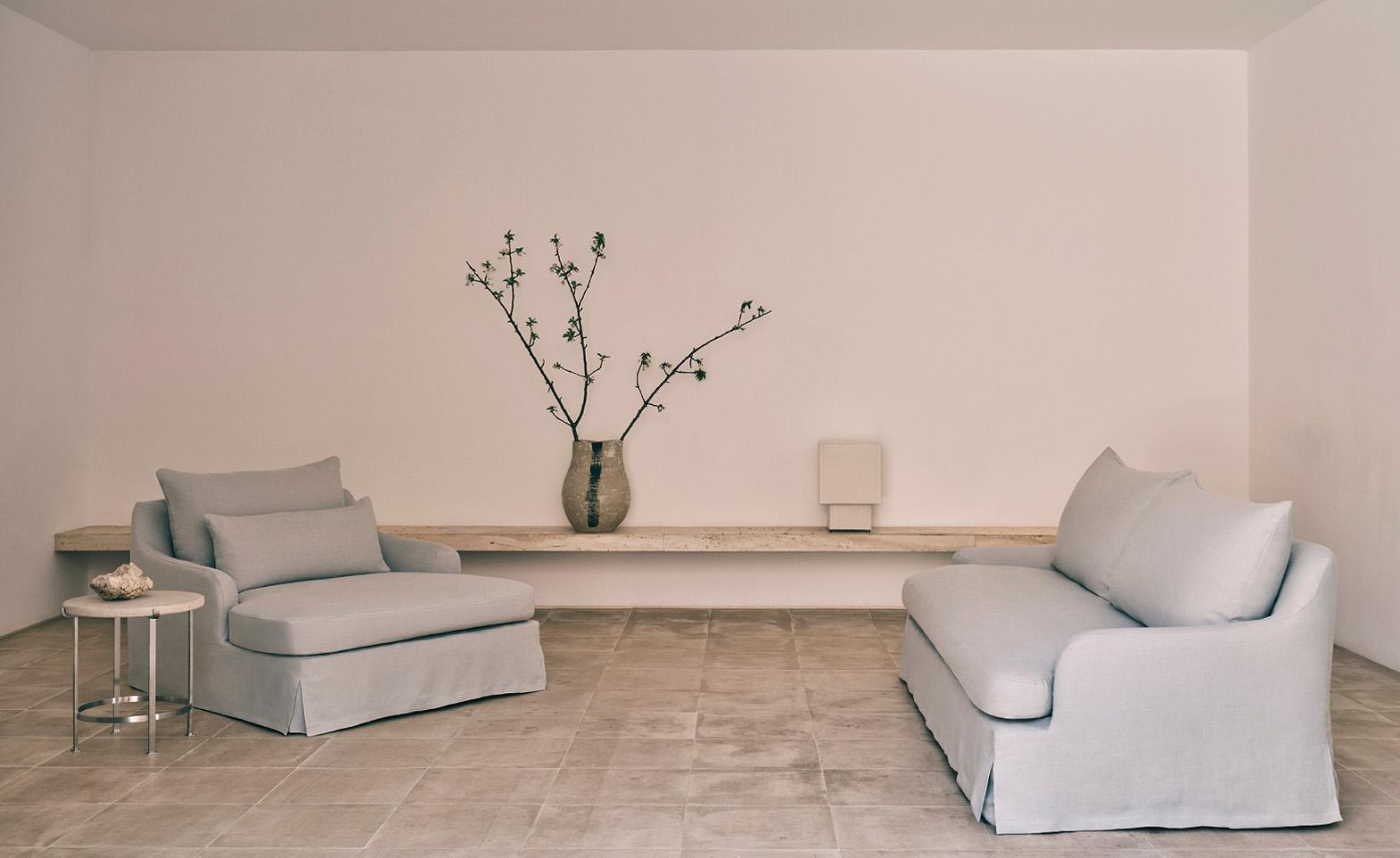 Vincent Van Duysen x Zara Home living room furniture, including a slipcovered sofa and arm chair, pictured alongside a circular side table, sculptural vase, and geometric table lamp