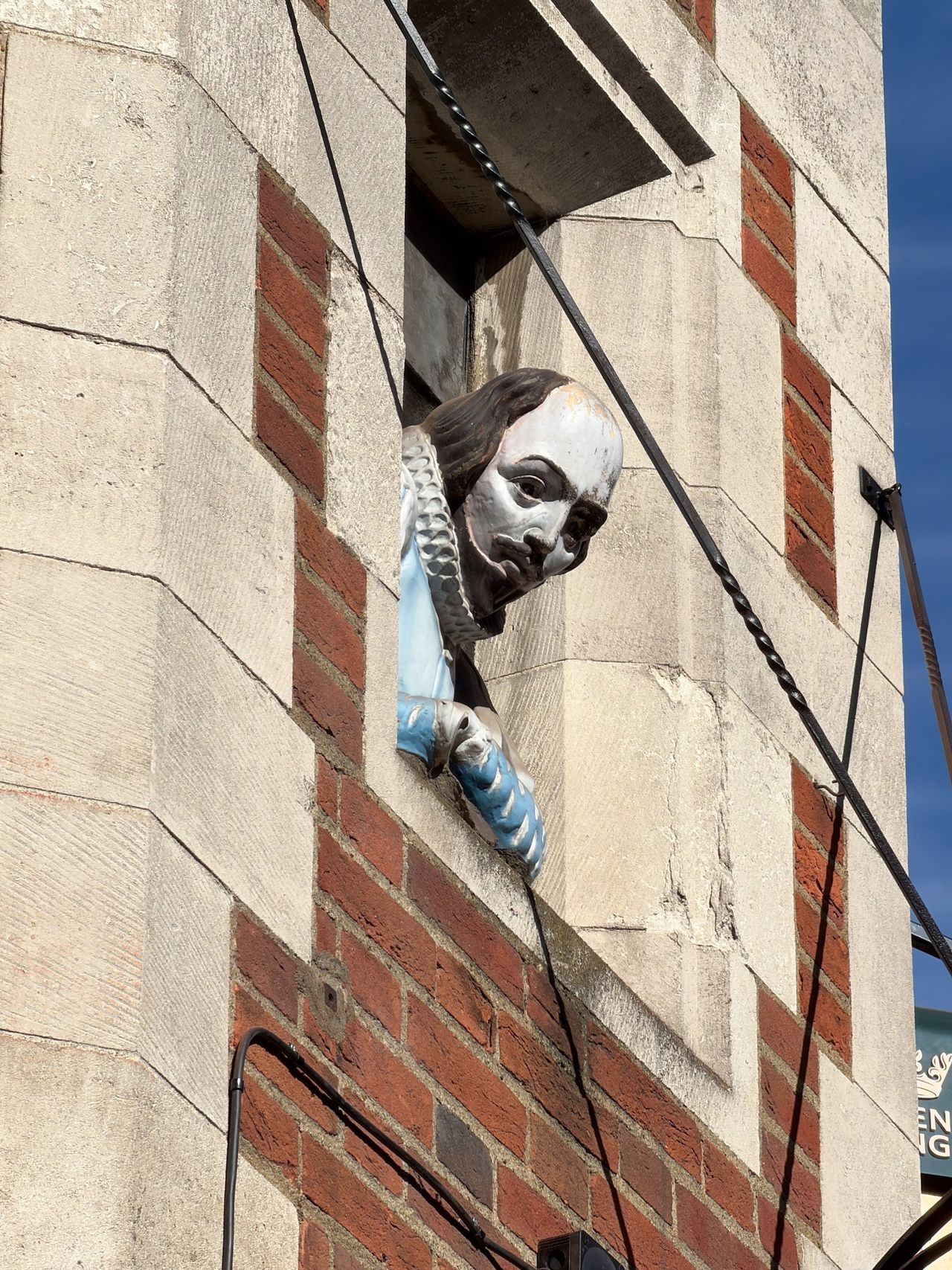 Painted figure resembling William Shakespeare leaning from a window in a red brick and stone building.