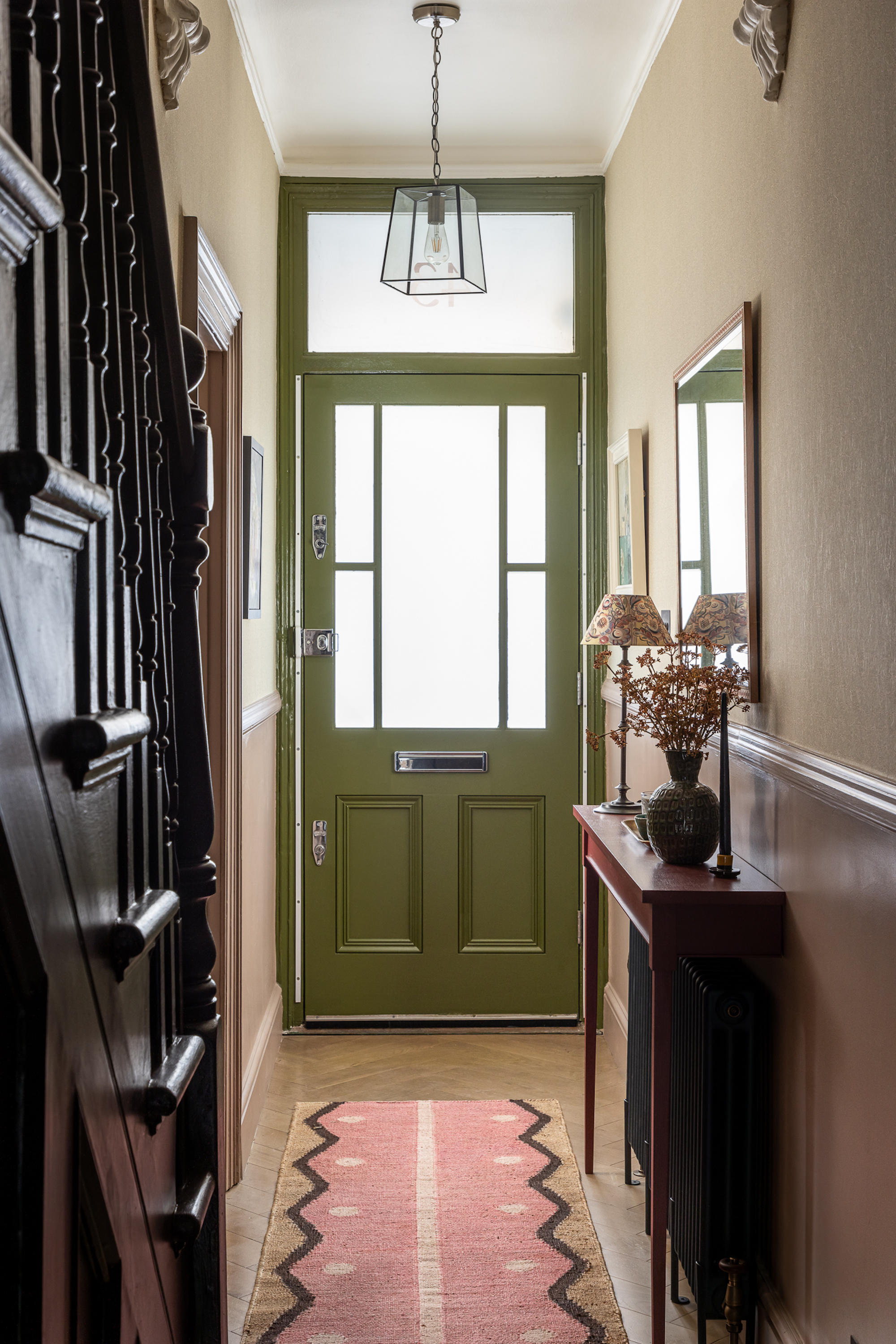 A hallway with a view of the front door, painted olive green. On the right is a small side table in a plumb red colour, and a staircase on the left in the same color.