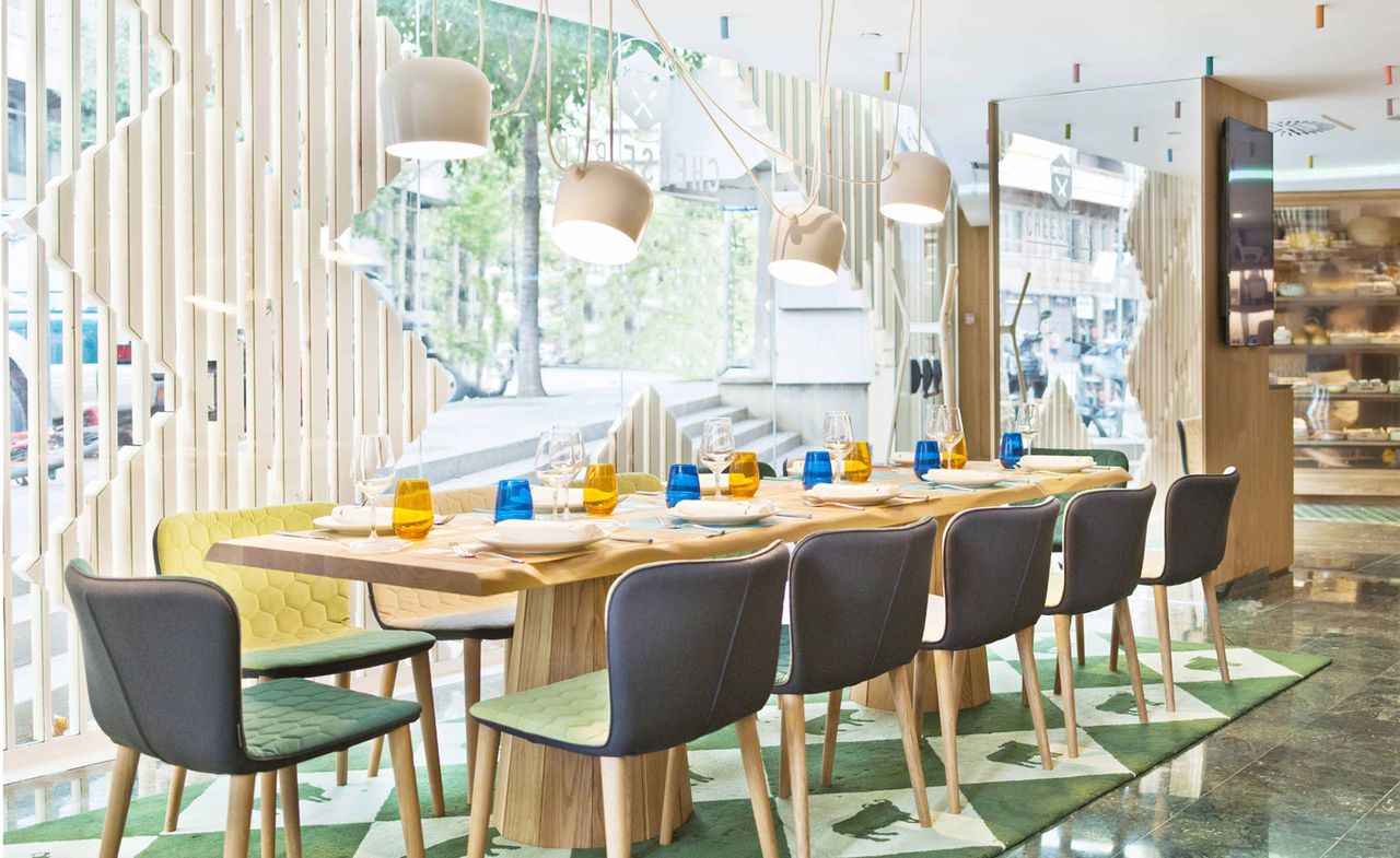 A restaurant with a long decorated table, blue chairs, patterned rug, a pendant light and a large window.