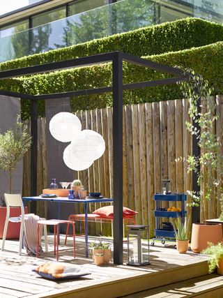 a deck with a modern black minimalist pergola with hedges beyond, and a child's red and blue dining set underneath