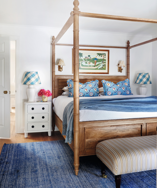 double four poster wooden bed with blue and white bedding and blue rug and ottoman and bedside table with blue check lamp