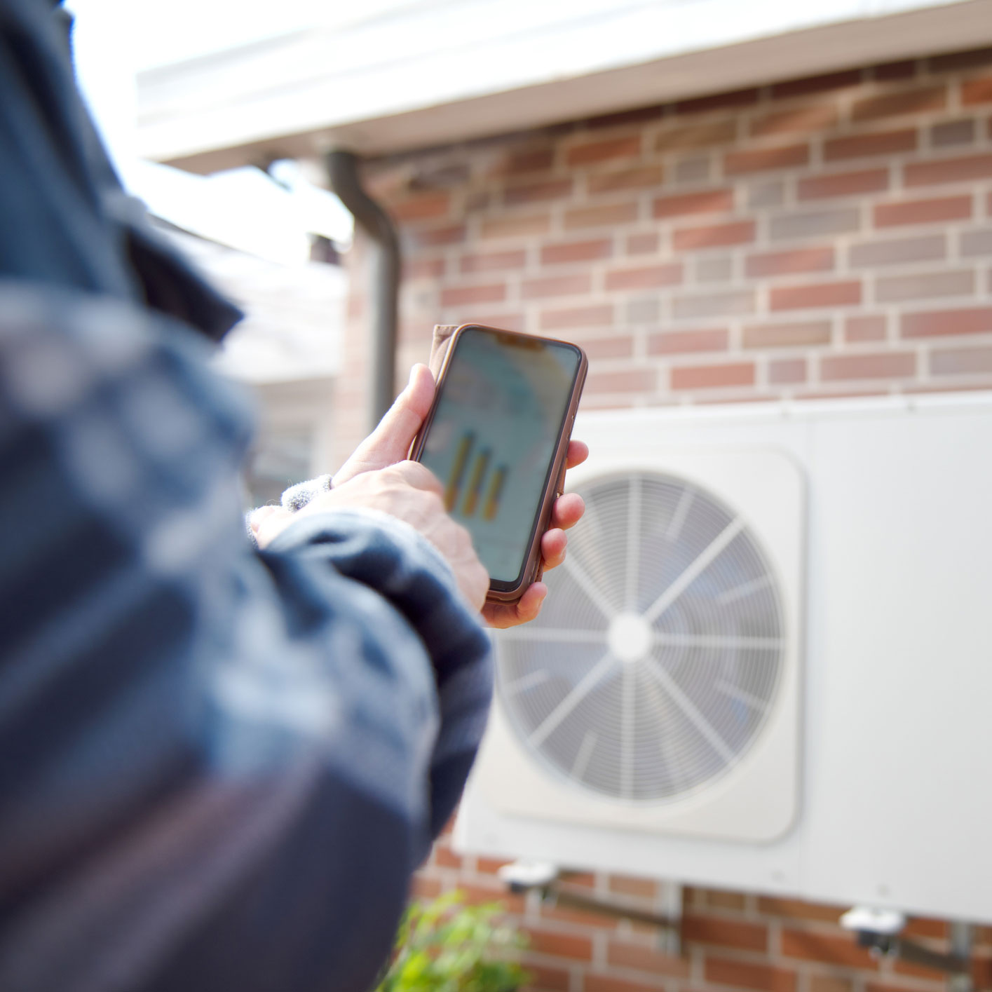 Arm holding a smart phone showing an app with a heat pump on a brick wall in the background