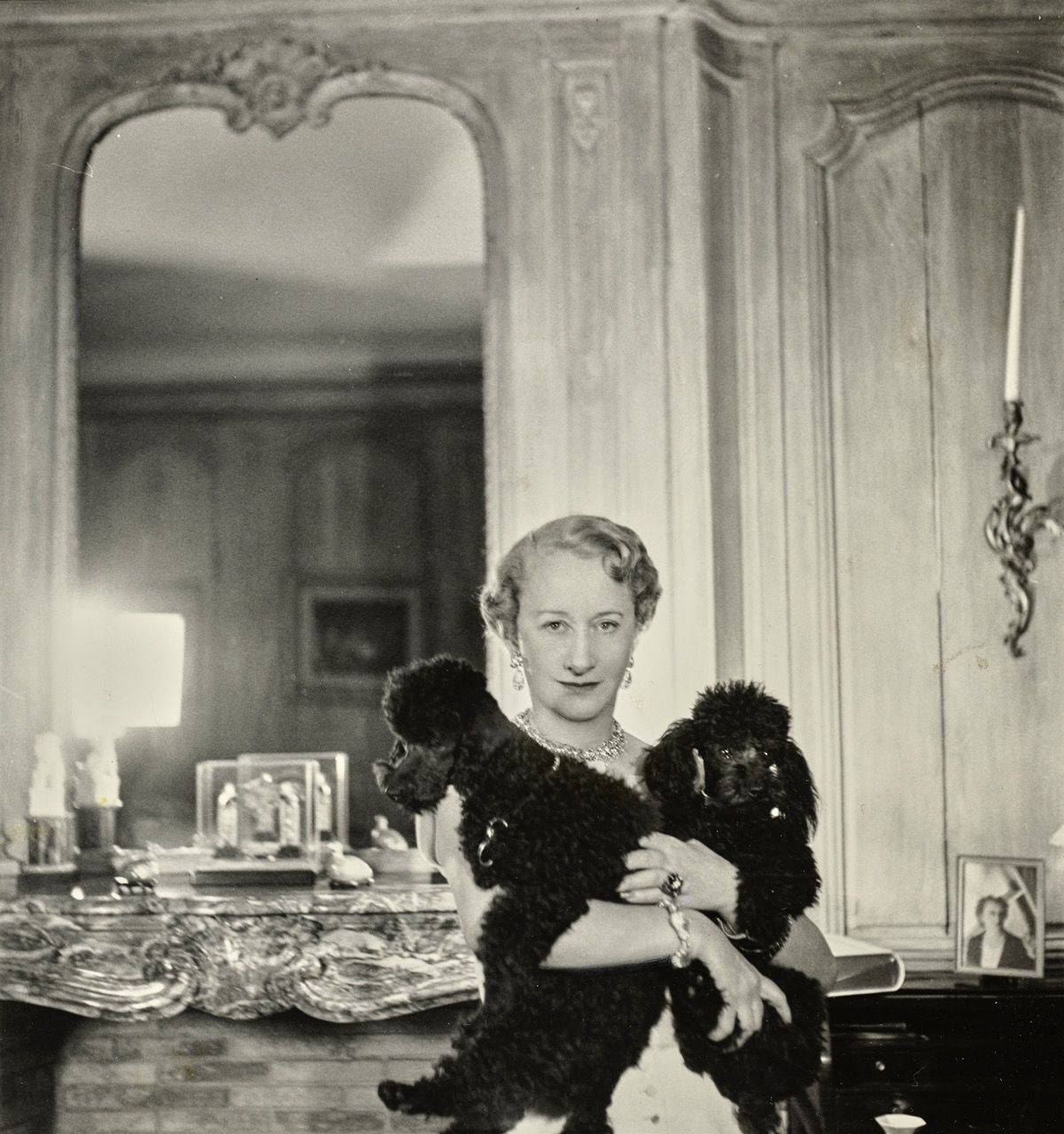 A woman wearing jewellery holds two black toy poodles inside an ornate interior.