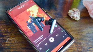 A pair of EarFun Air Pro 3 wireless earbuds sitting atop a Google Pixel 7 Pro, the phone's screen showing Overcompensate by Twenty One Pilots playing on Spotify.