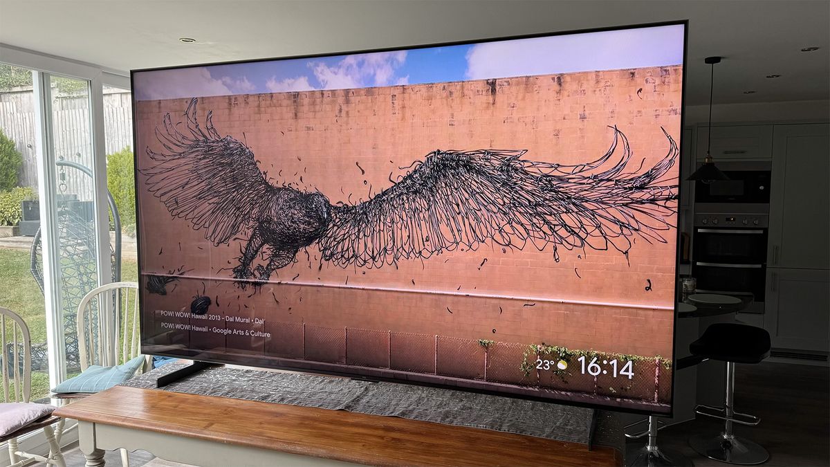 The TCL 98C7K 98-inch TV on a table in a living area, seen at a slight angle. On screen is a brick wall featuring an illustration of a bird of prey.