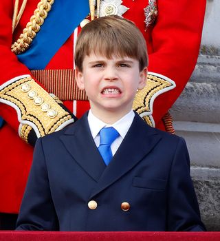 Prince Louis wearing a blue suit and tie gritting his teeth while standing in front of Prince William, wearing a red military uniform, on the balcony at Buckingham Palace