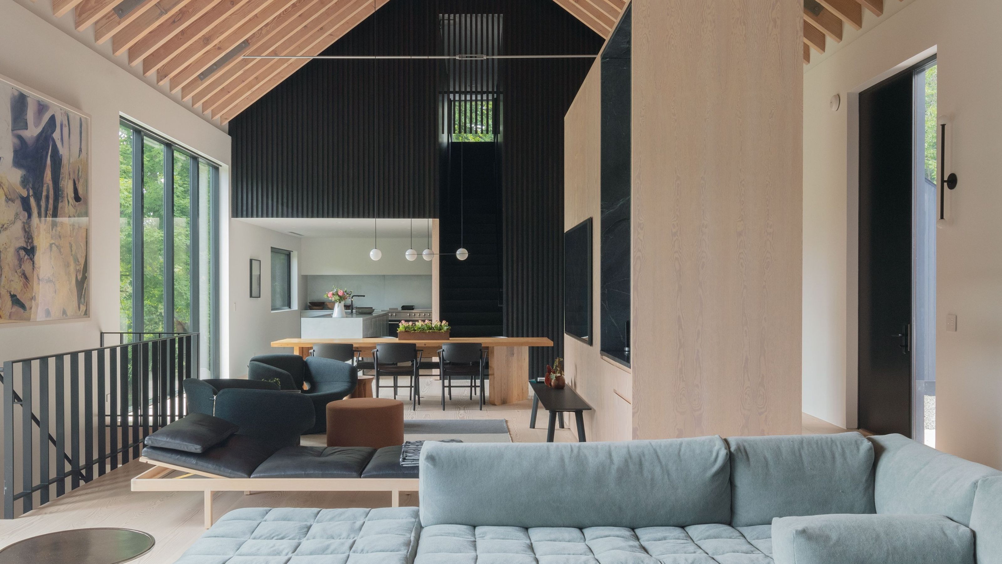 modern home with raked ceiling timber panelling, black timber slats, light blue sofa, with timber table and armchairs and black stair balustrade going downstairs