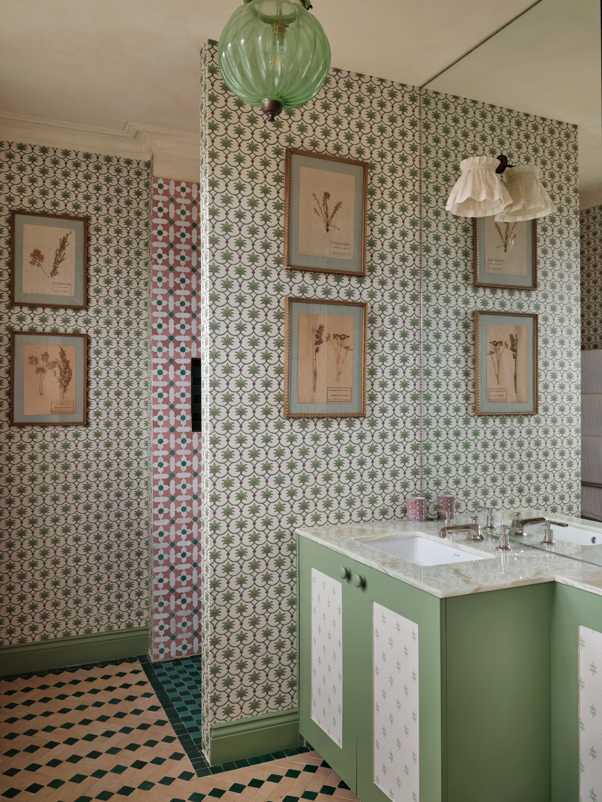 A maximalist bathroom with vintage framed artwork in botanical motifs, and a plethora of patterned tiles in different styles and colors, features a mint green basin topped with marble.