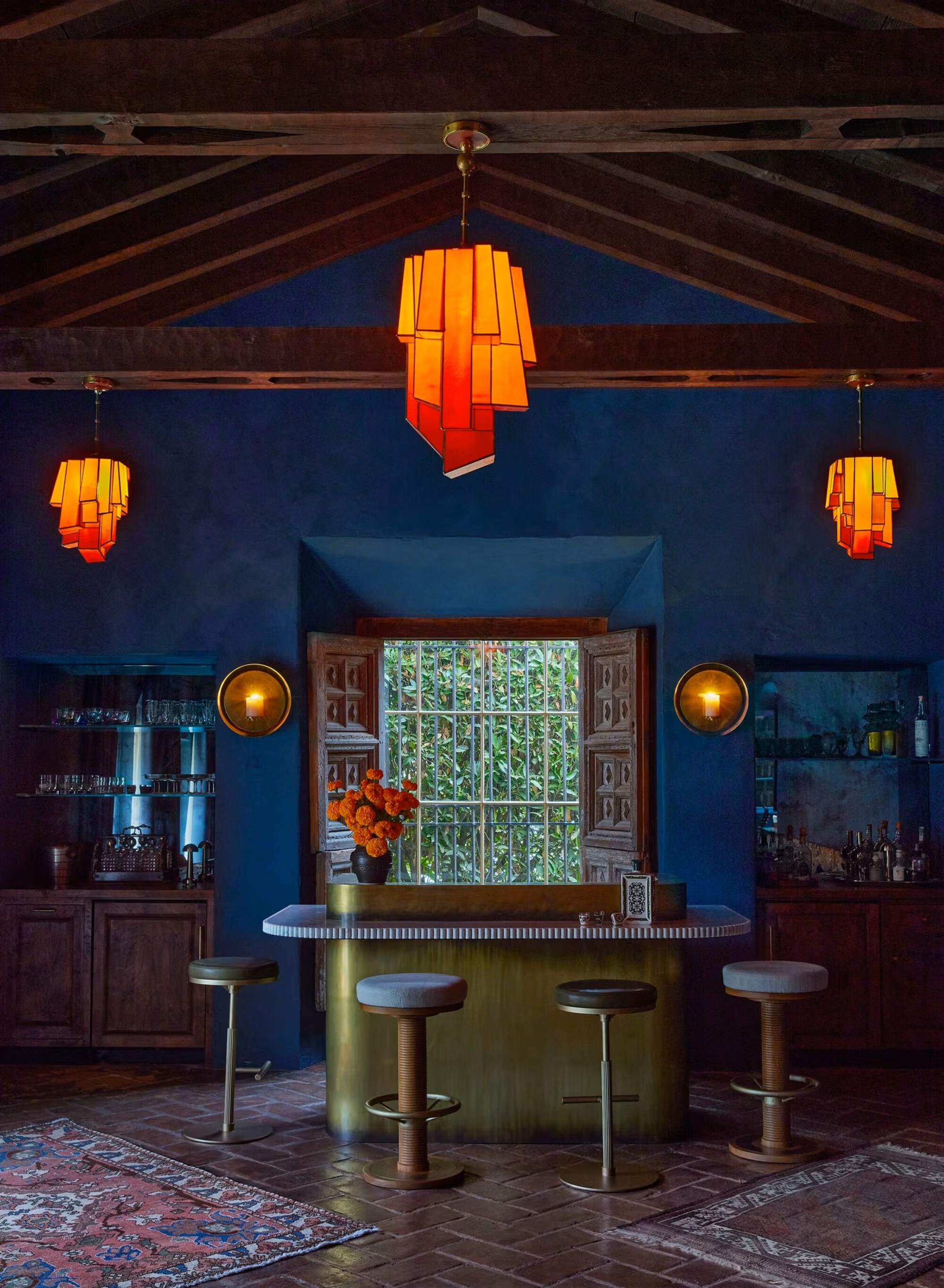 A home bar room with dark blue walls, wooden ceilings, and brick flooring. There is a gold bar, three orange pendant lights, and two rugs in the room.