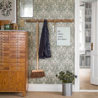 a wallpapered hallway with a large storage unti til one side and a wooden coat rail on the wall with a sweeping brush and coat hanging from it