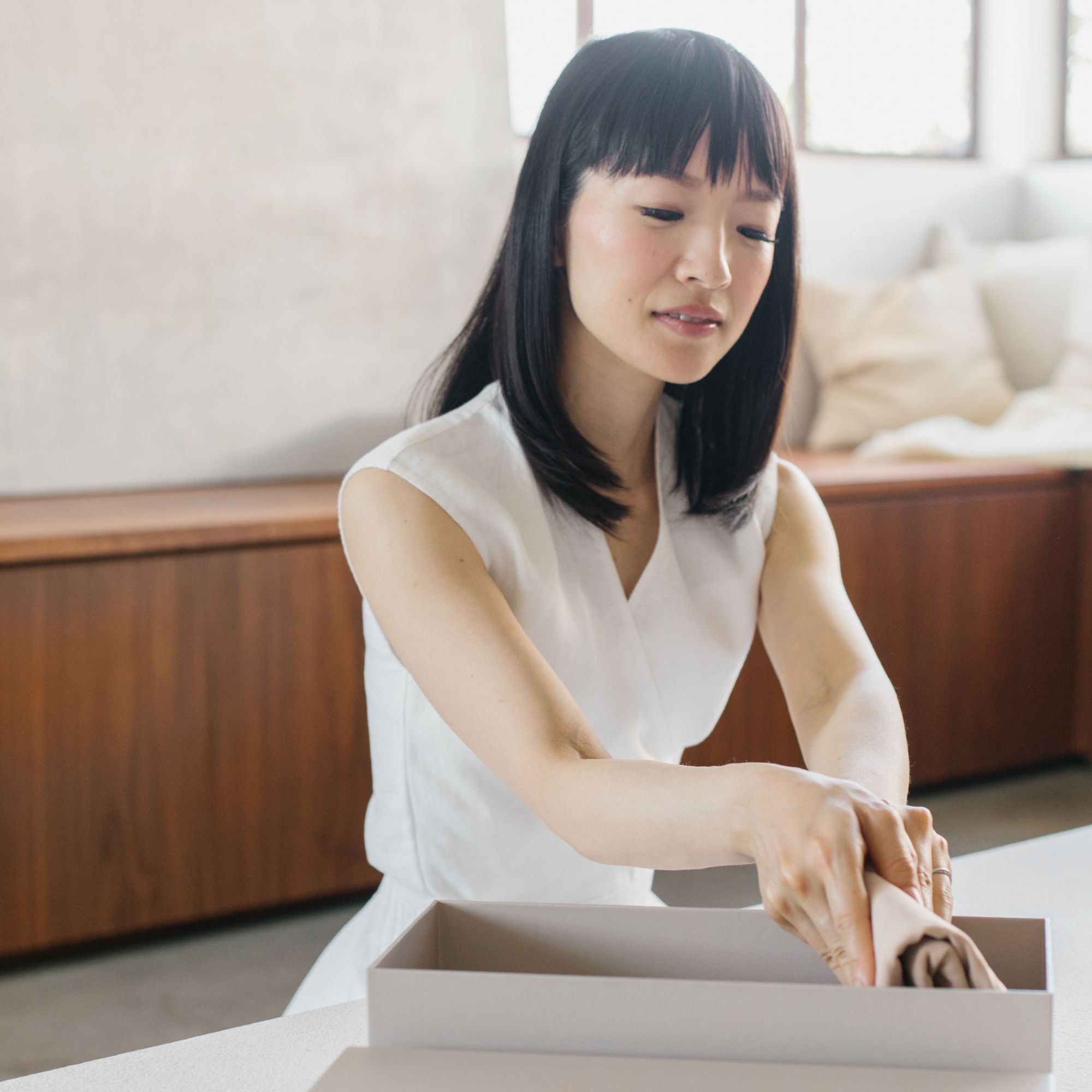 Marie Kondo wearing a white shirt, sitting on the floor and folding clothes in a beige clothes organiser