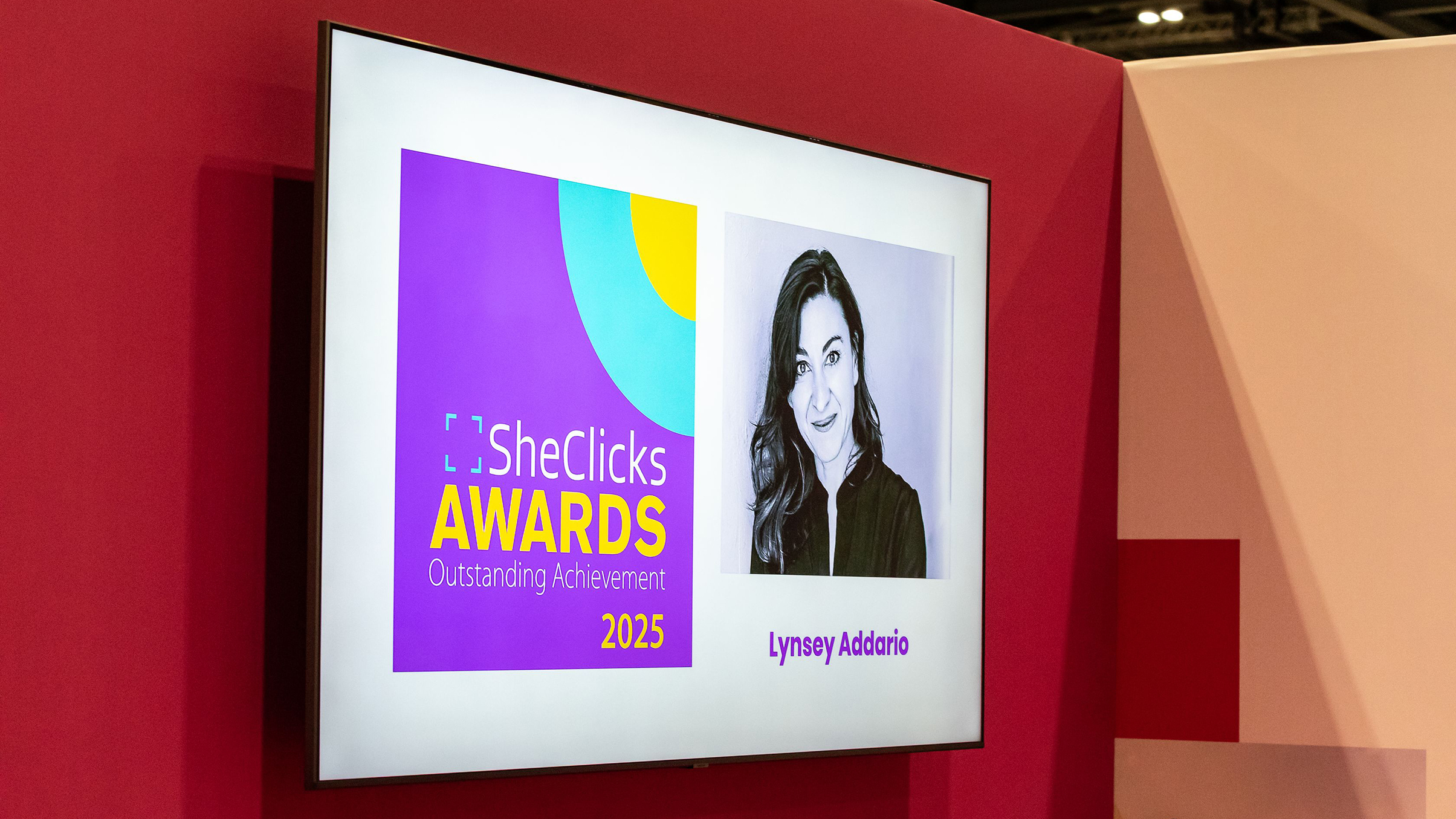 A digital display announces the SheClicks Awards 2025, featuring an image and the name "Lynsey Addario" on a vibrant purple background