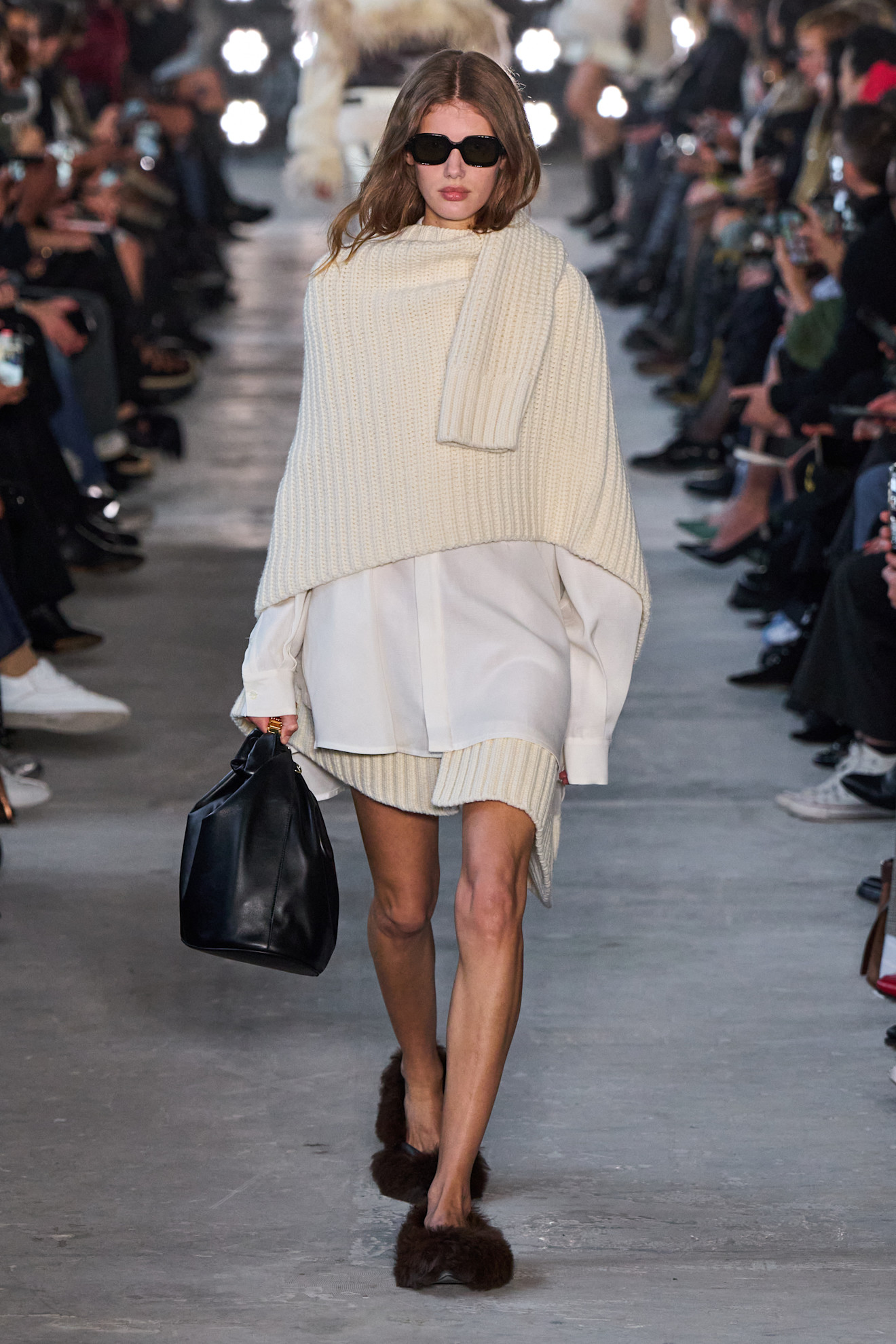 Fur accessory trend is shown in Sacai&#039;s fall-winter 2025 collection in a close photo of a model walking down the runway wearing a white shirt dress with a sweater layered on top like a cape and a mini knitted skirt underneath, styled with black sunglasses, a black hobo nah, and brown furry flats