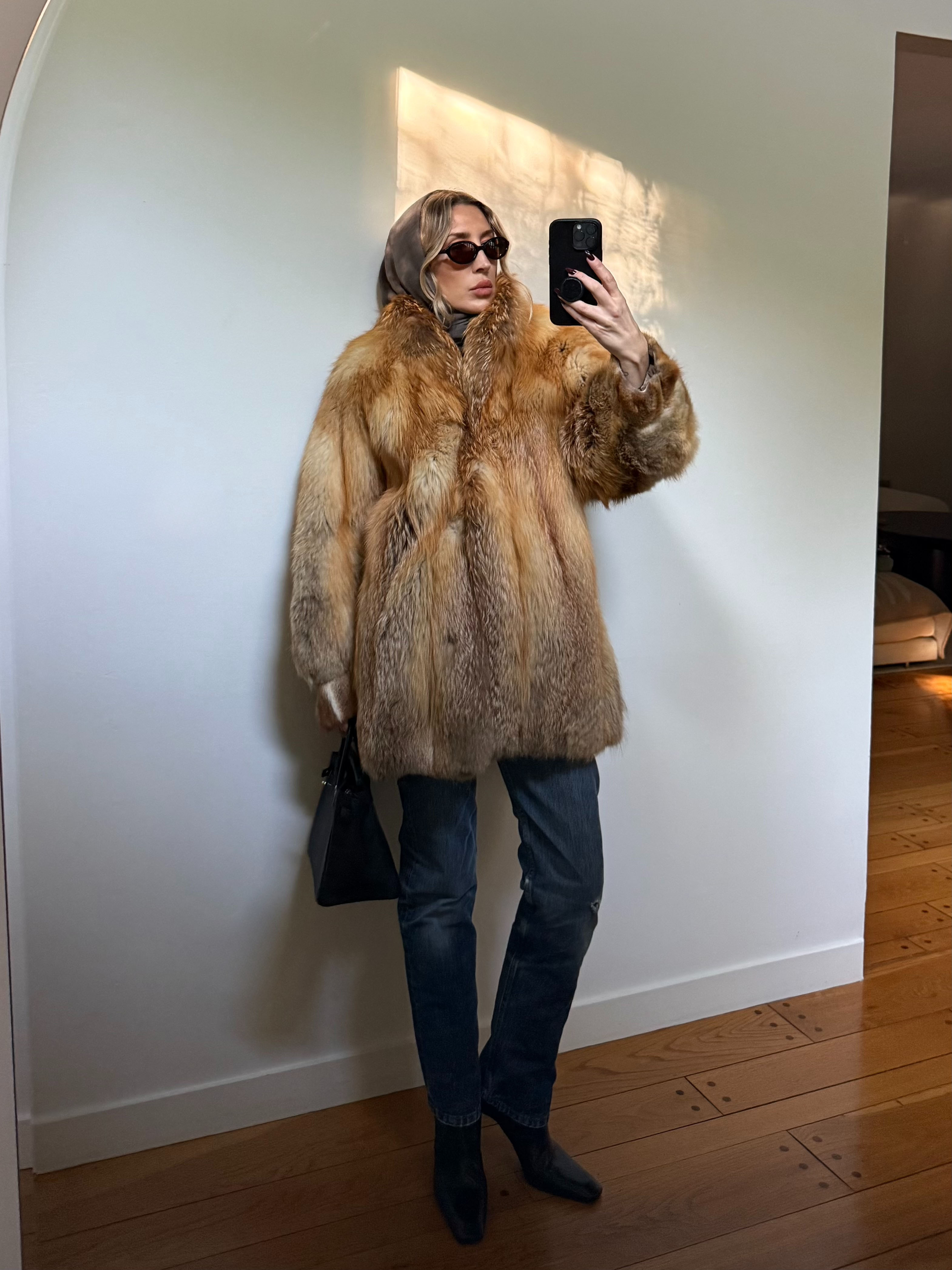 Woman taking a selfie in an amber fur coat, dark denim jeans, oval sungalsses, black small tote bag and iPhone