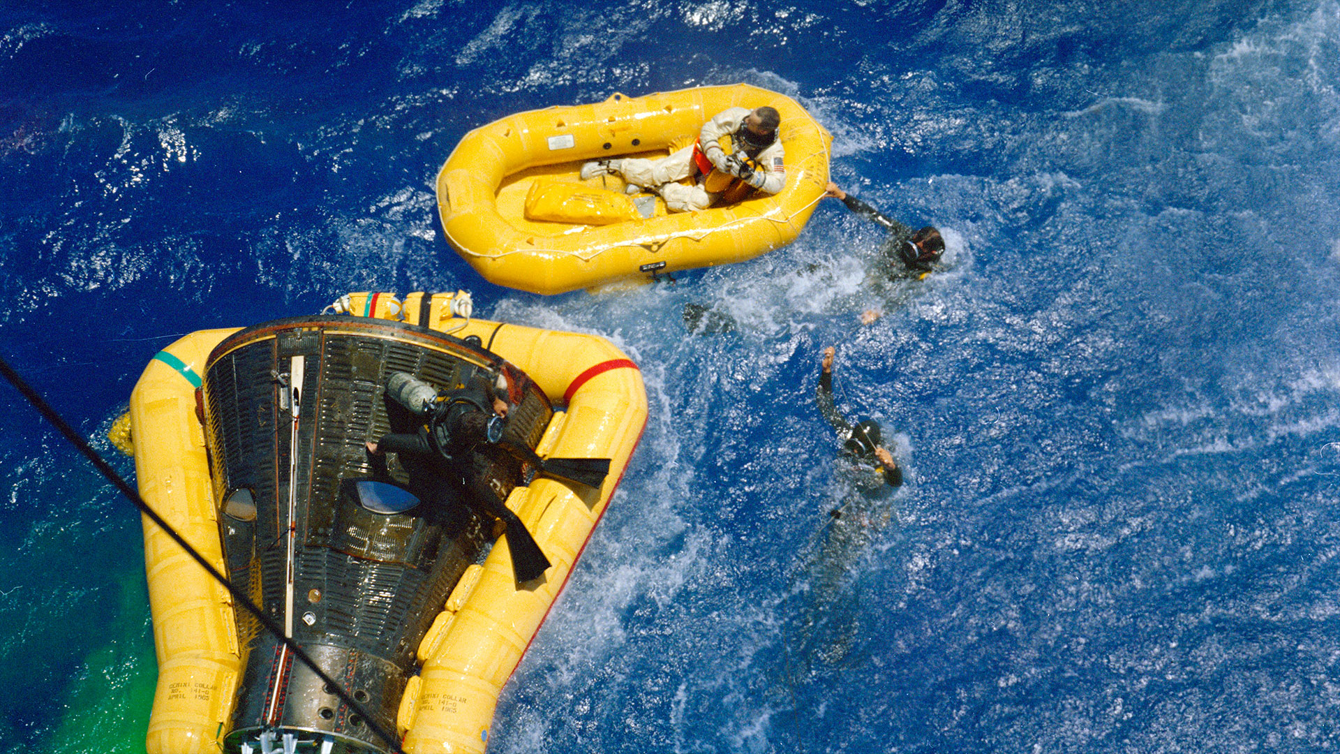 a bright yellow yellow holds a man in a spacesuit while other men in the deep blue blue work around a space capsule surrounded by a similar yellow floatation collar.