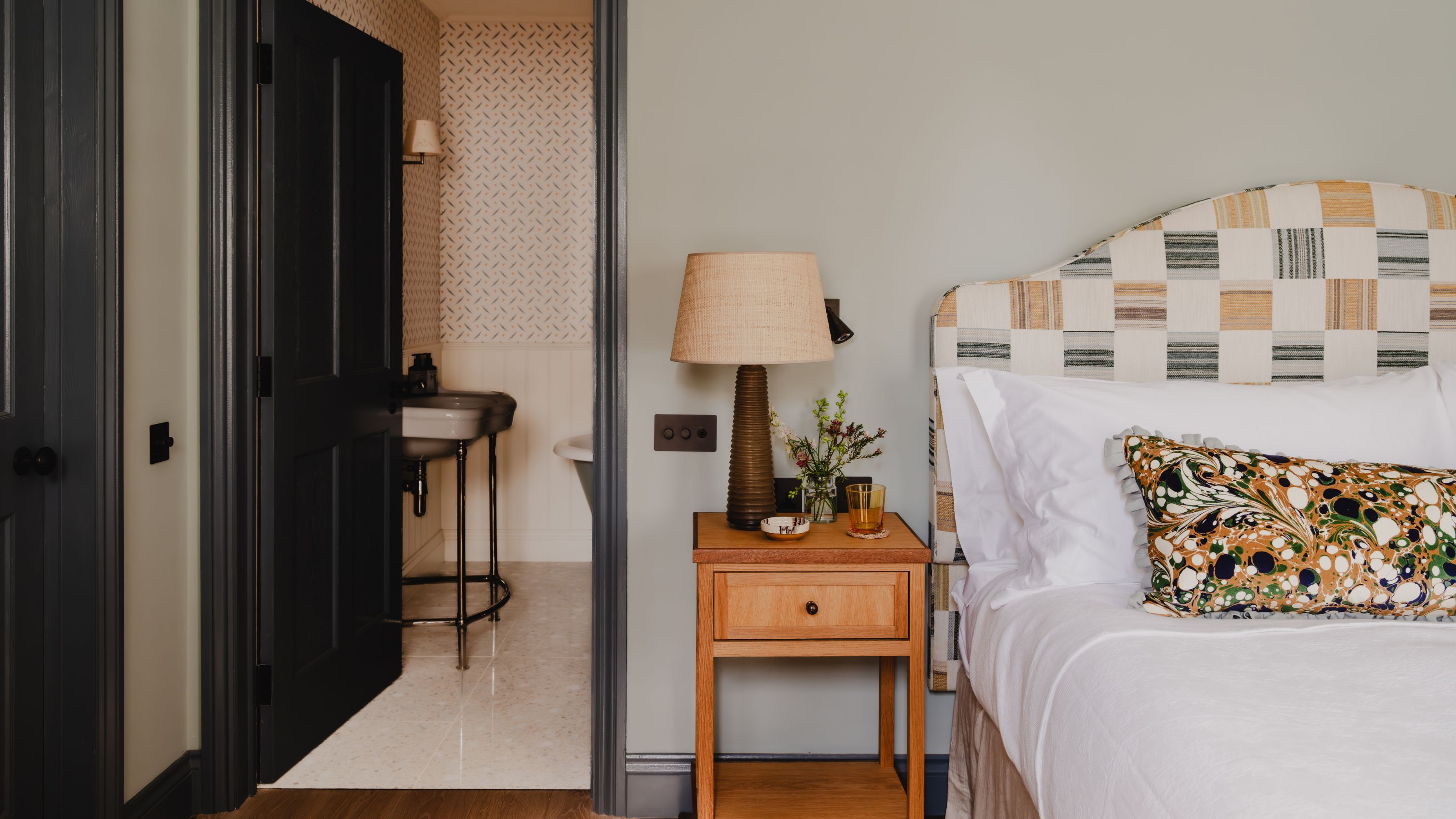 a pale blue bedroom with navy wood trim and doors with a view into a wallpapered bathroom. the bed has a yellow and green patchwork headboard