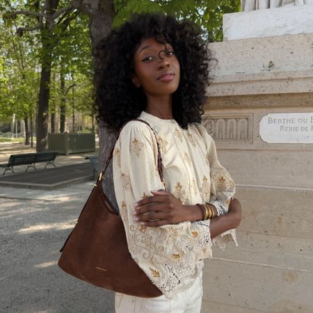 Woman wears embroidered blouse, white jeans and carries brown suede bag