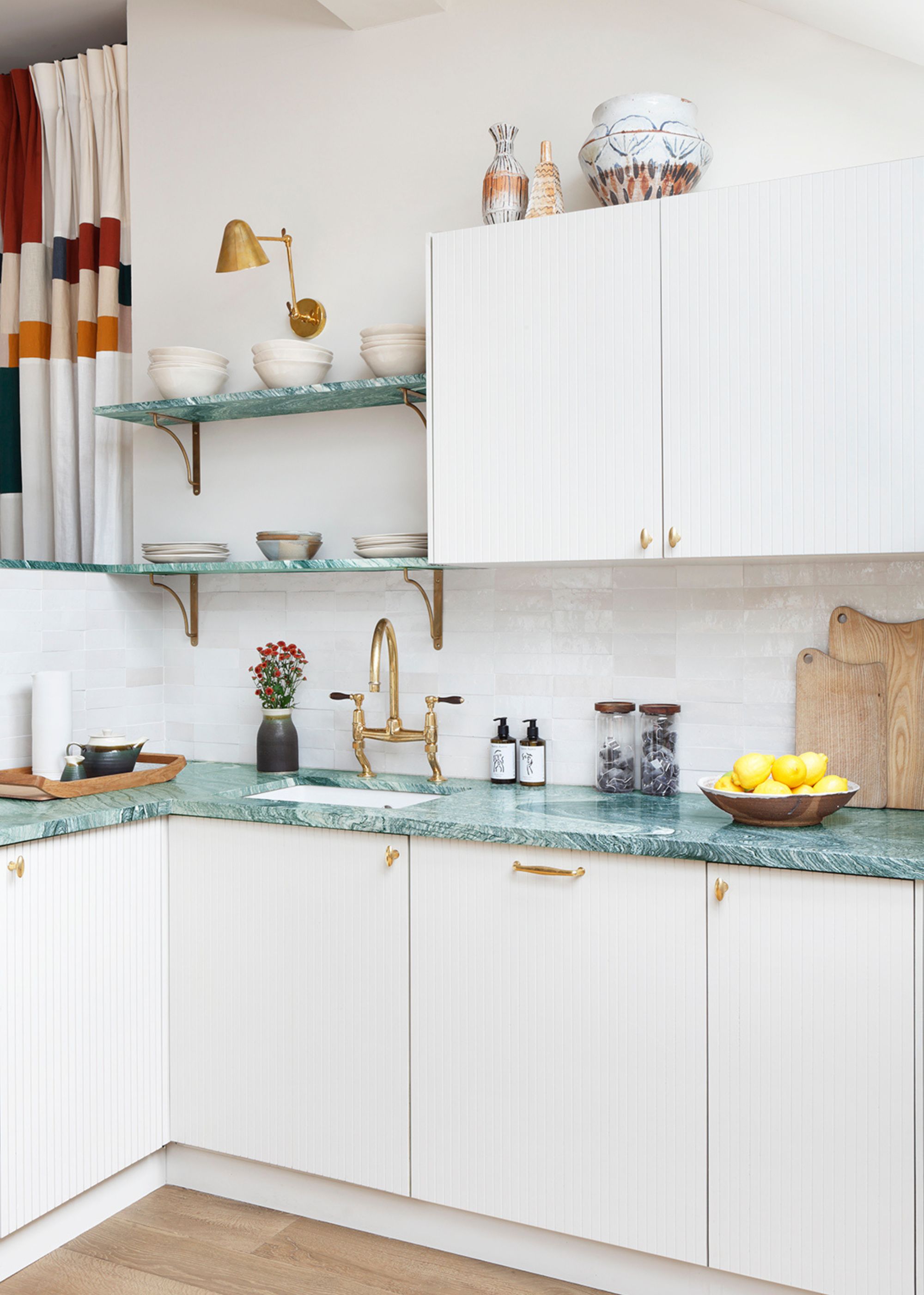 A modern kitchen with white cabinets and walls, green stone countertops, open shelving, brass handles, light fixtures, and a tap.