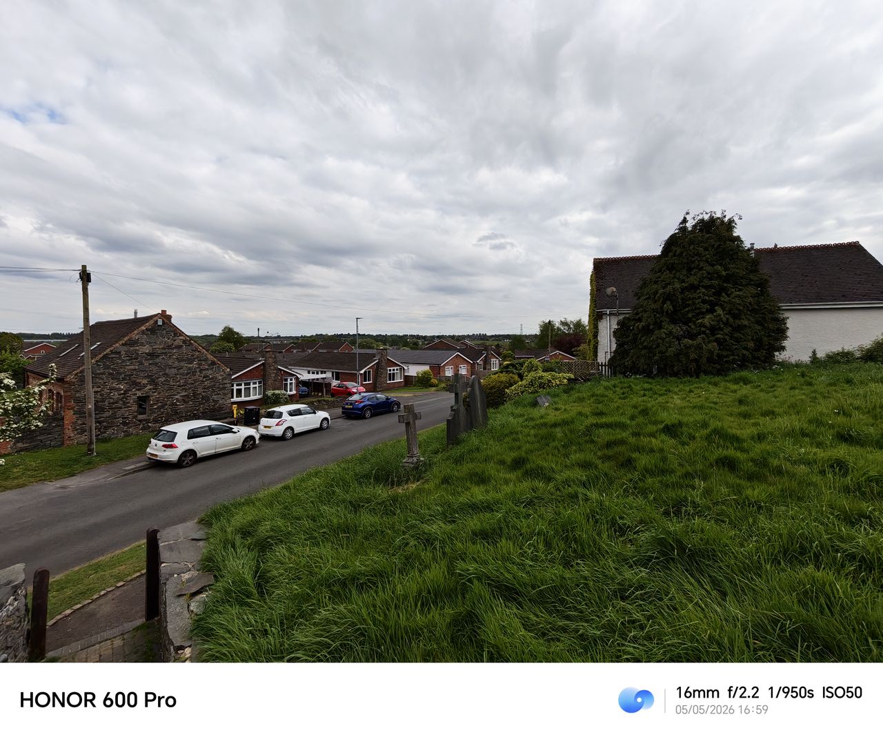 Wide-angle view of a village street and grassy churchyard under a cloudy sky