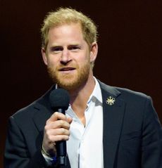 Prince Harry wearing a blue suit speaking into a mic in front of a dark background