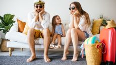 Family wearing sunglasses sitting on the couch next to their suitcases looking sad because of canceled trip