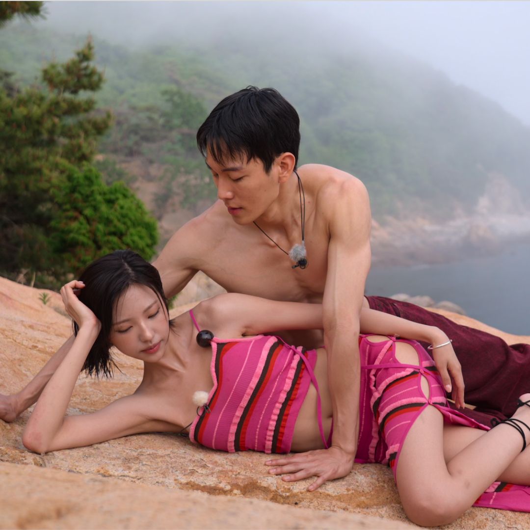 A woman wearing a pink striped cutout gown (Kim Go-eun) props her head on her hand as she lies on a large rock. A shirtless man wearing red pants (Kim Jae-jin) looks down at her, leaning on his hands, in a still from 'Single's Inferno' season 5.
